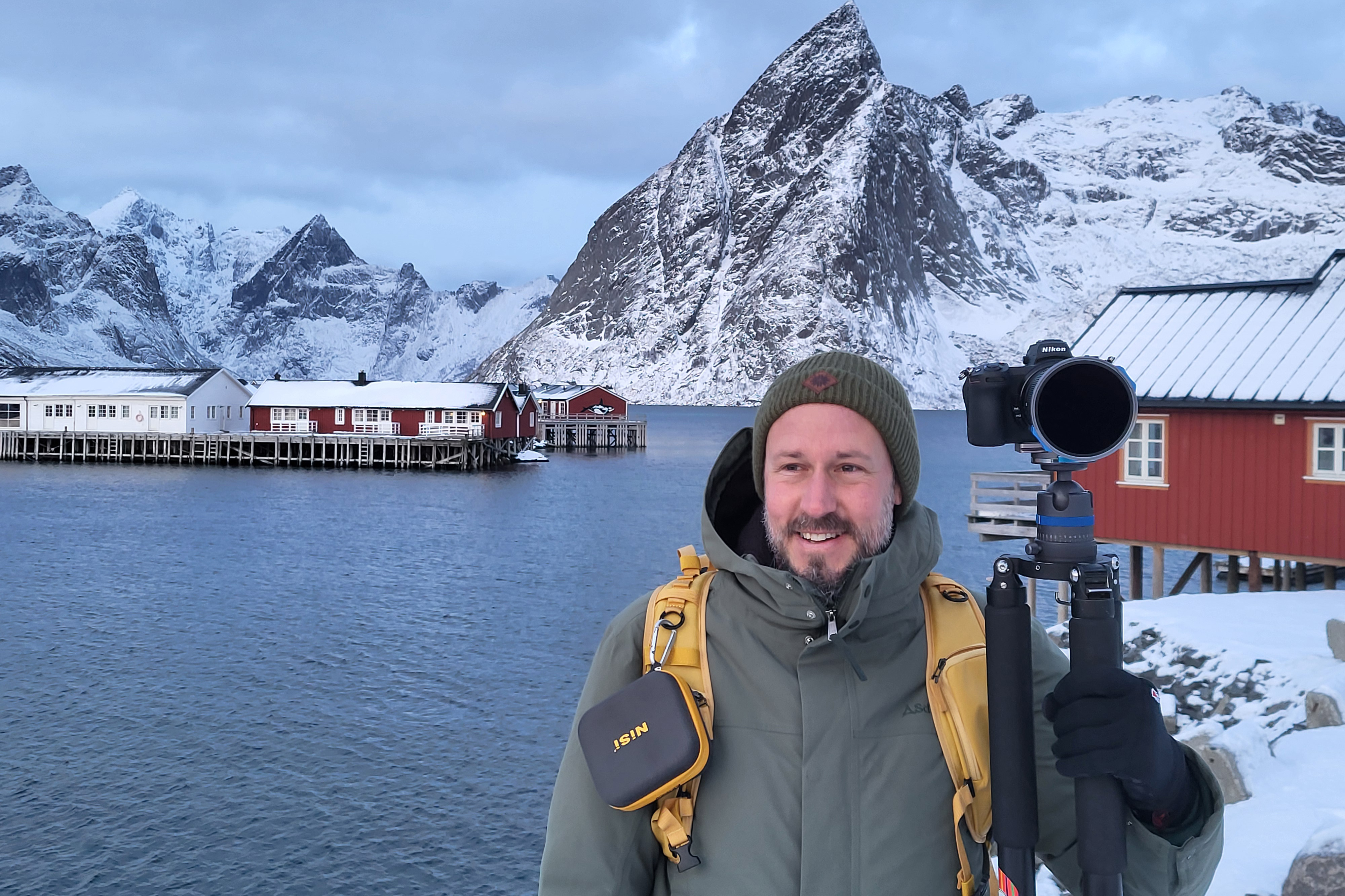  Photographer standing in snowy Lofoten landscape with camera on tripod, red cabins and dramatic mountains 