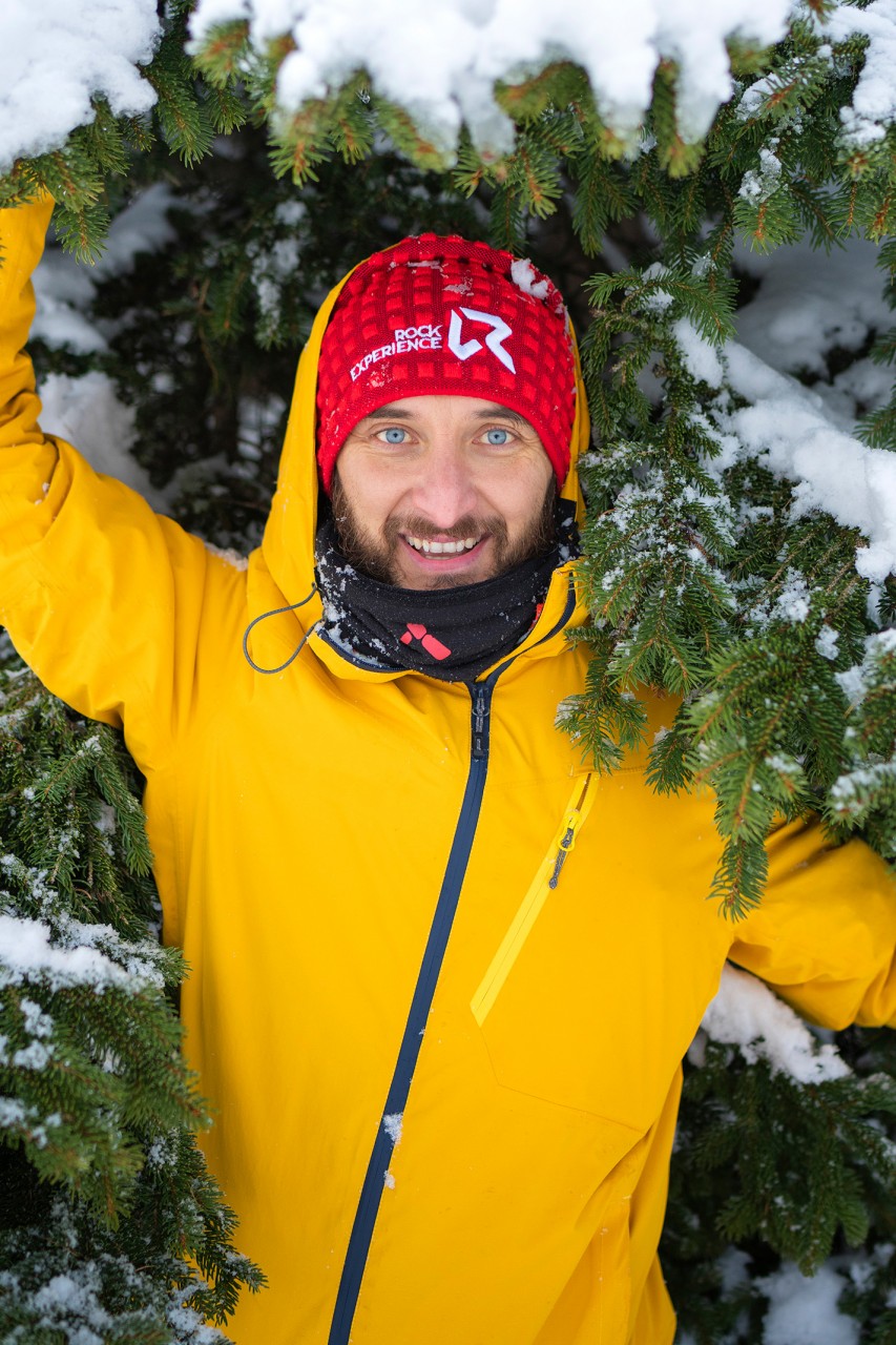 Homme aux yeux bleus, bonnet et veste d'hiver devant un arbre enneigé