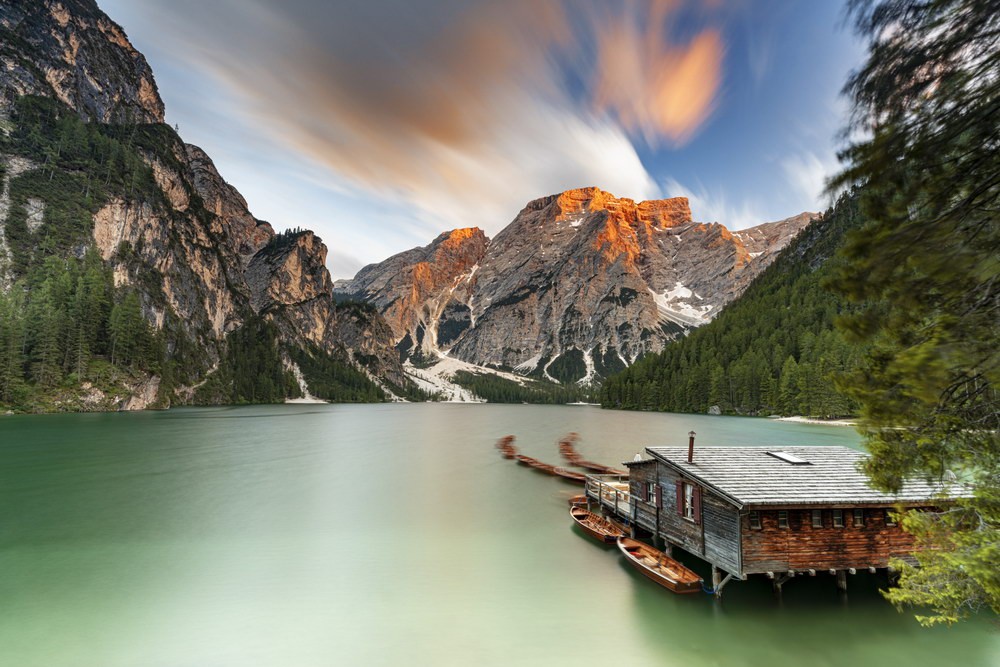 A boat hut with fishing boats on the green Braies Lake and mountains in the background.