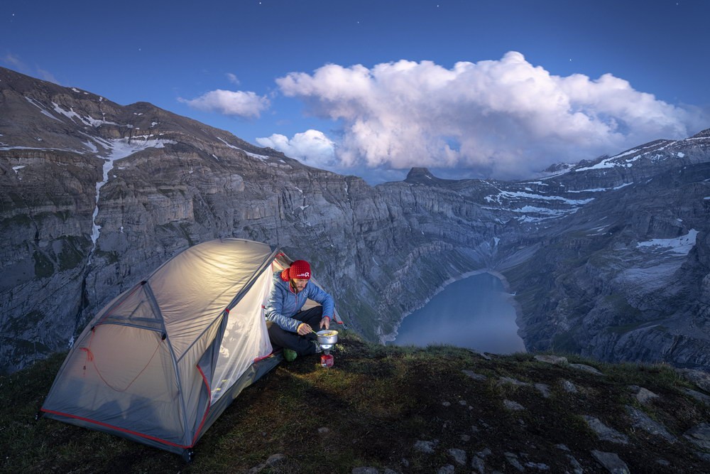 Man with camping cooker in front of tent amidst a mountain backdrop with lake