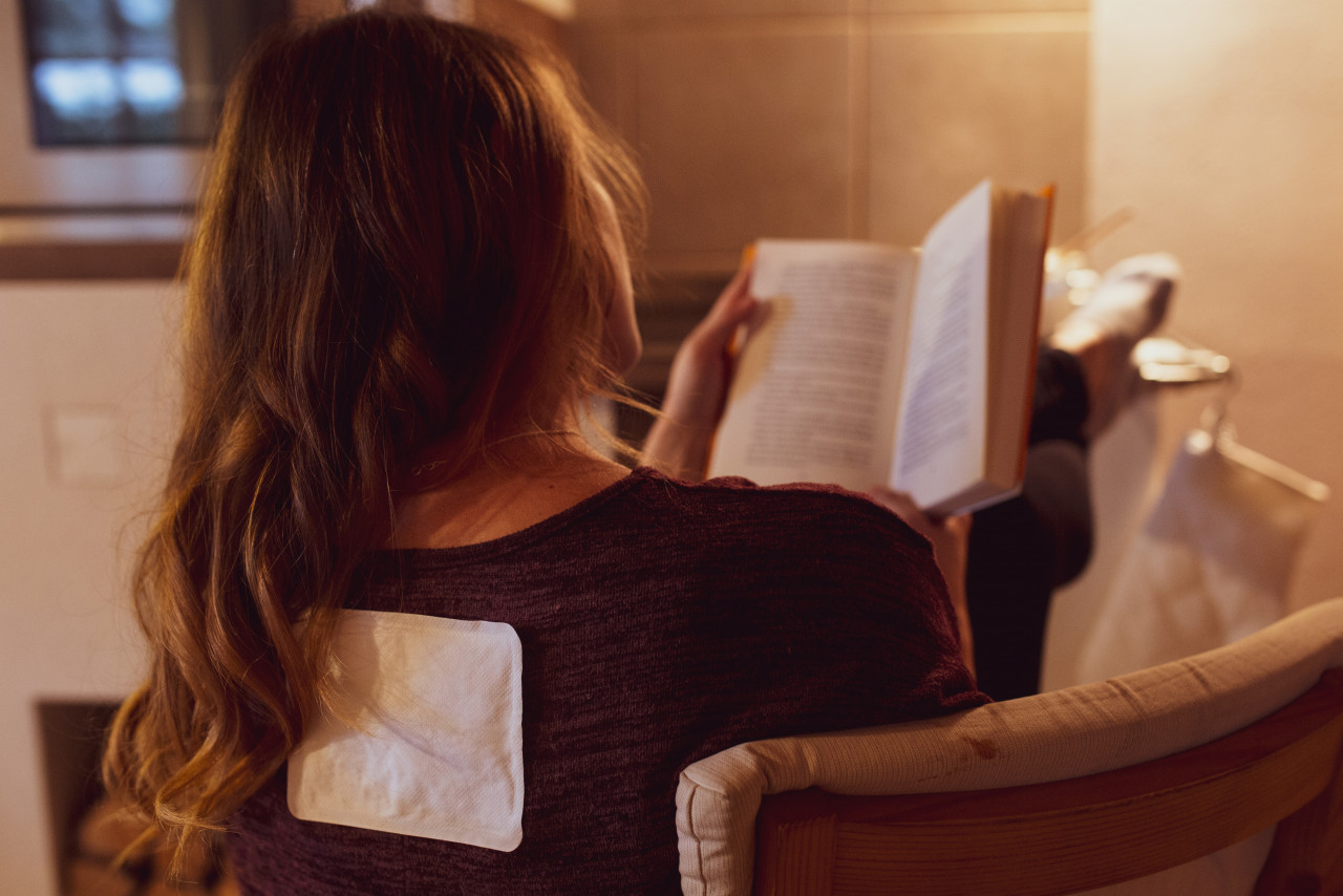 Mujer sentada en un sillón leyendo un libro mientras lleva un calentador en la espalda