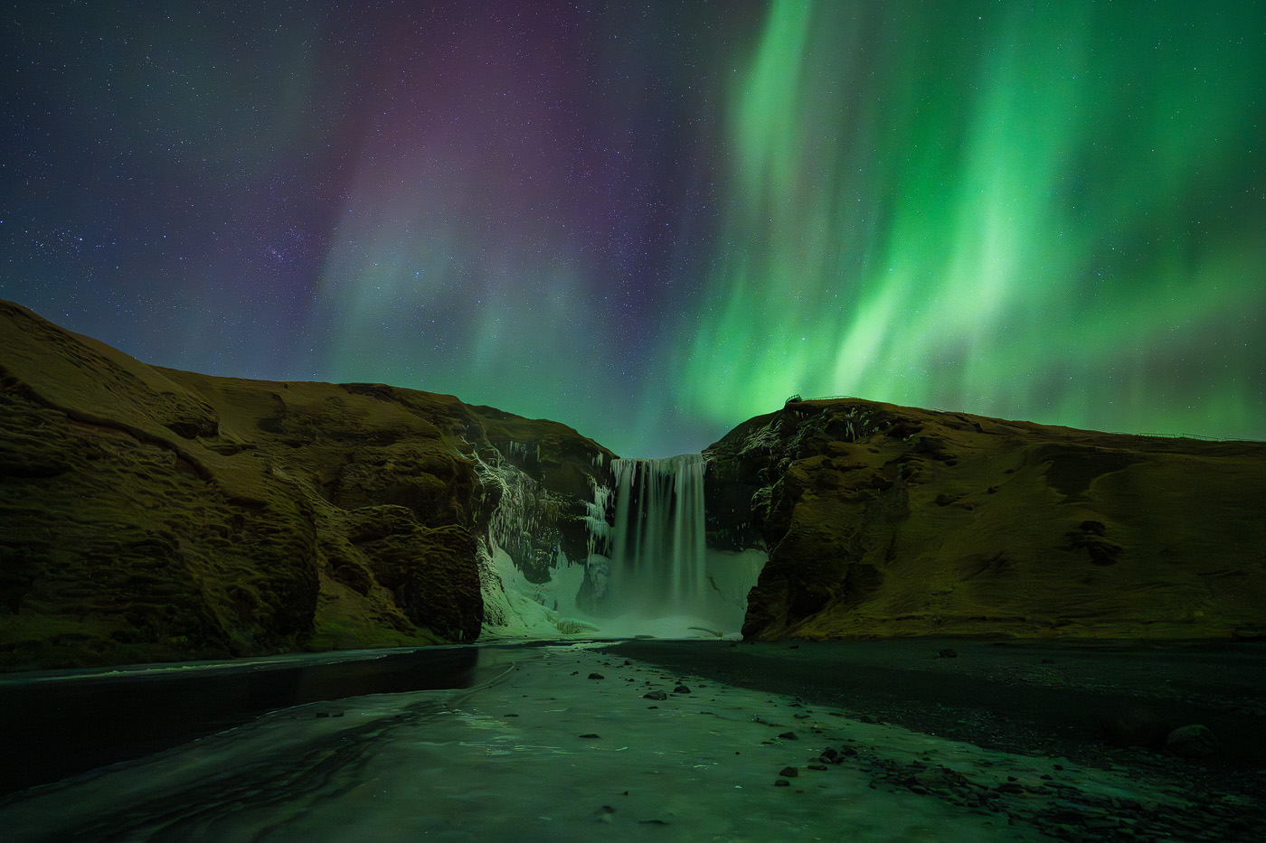 Green northern lights above a frozen waterfall in Iceland with reflections in an icy river at night.