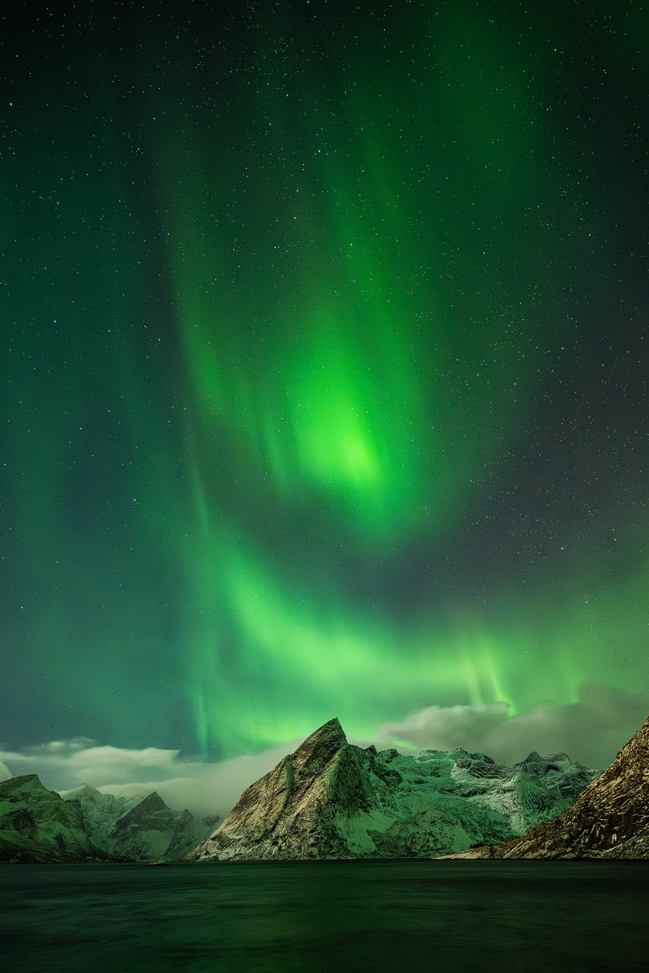 Green northern lights above snow-covered mountains and calm water in the Lofoten at night