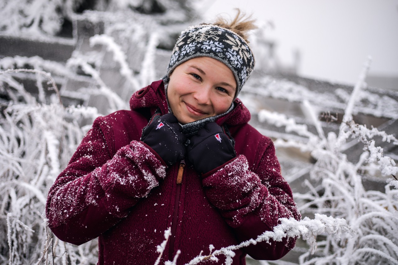 Lächelnde Frau mit Handschuhen, warmer Jacke und Stirnband inmitten von hohem Gras mit Raureif