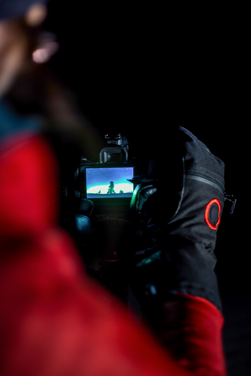Mensch mit Fäustlingen hält eine Kamera mit Live-Bild von einem Baum mit Polarlicht 
