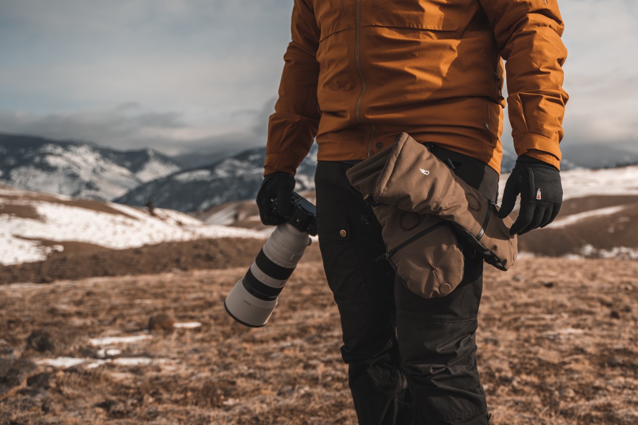 person in the mountains is holding a camera with gloves and has the mittens attached to the side of the pants with a carabiner