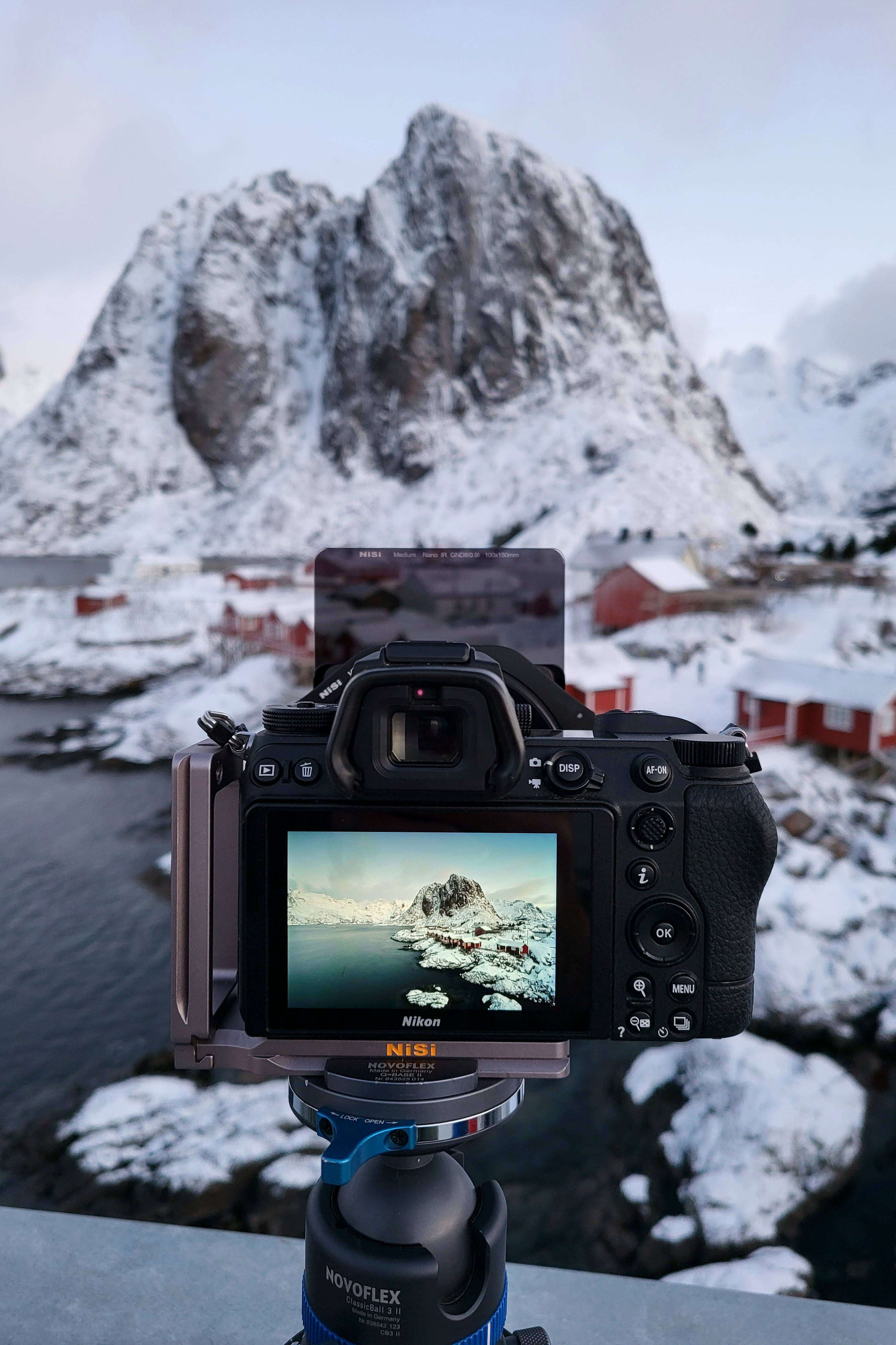 Camera on tripod with ND filter capturing snowy mountains and red cabins in the Lofoten Islands, Norway.