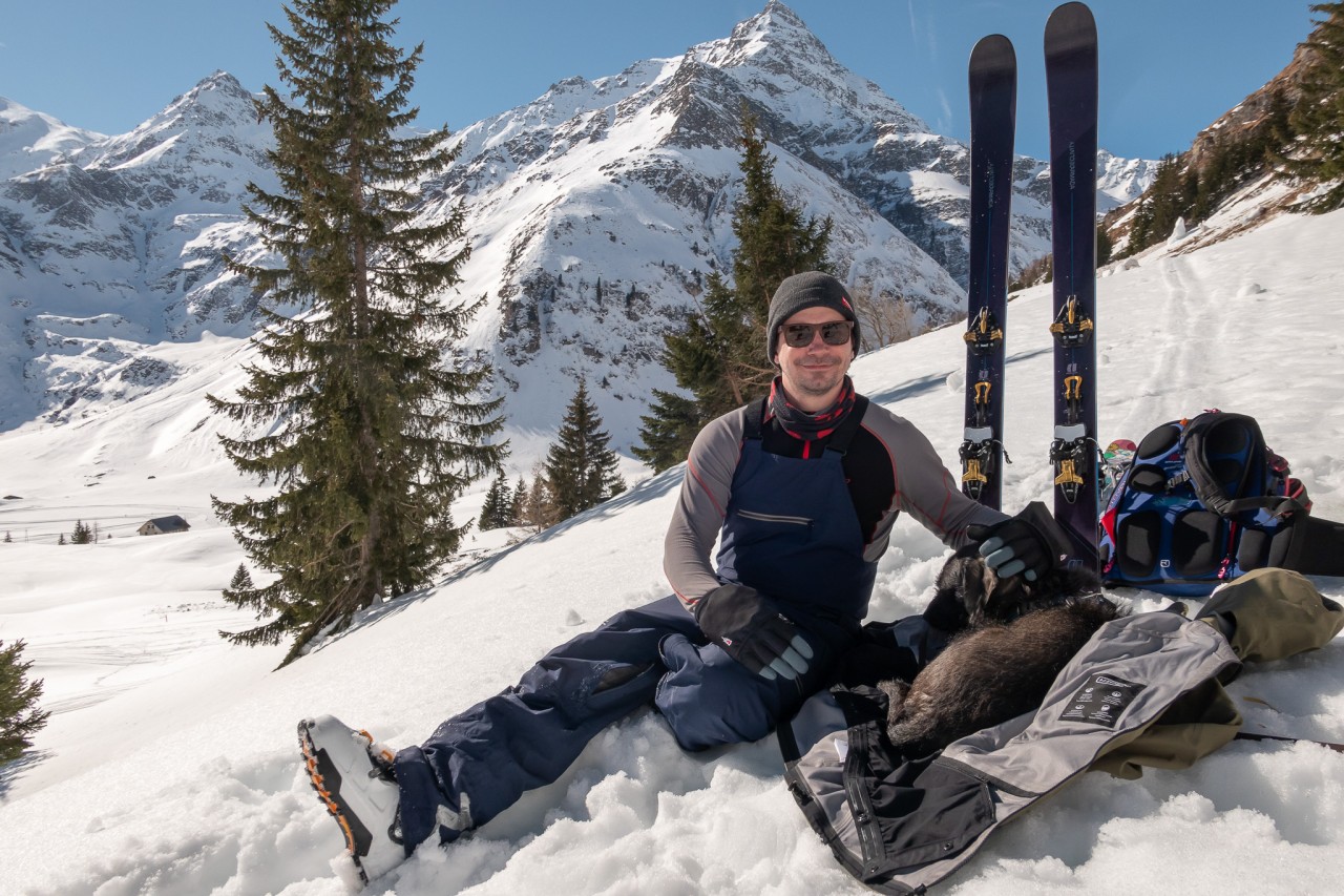 Uomo seduto sulla neve con il suo cane in montagna, con gli sci sistemati dietro di lui nella neve 