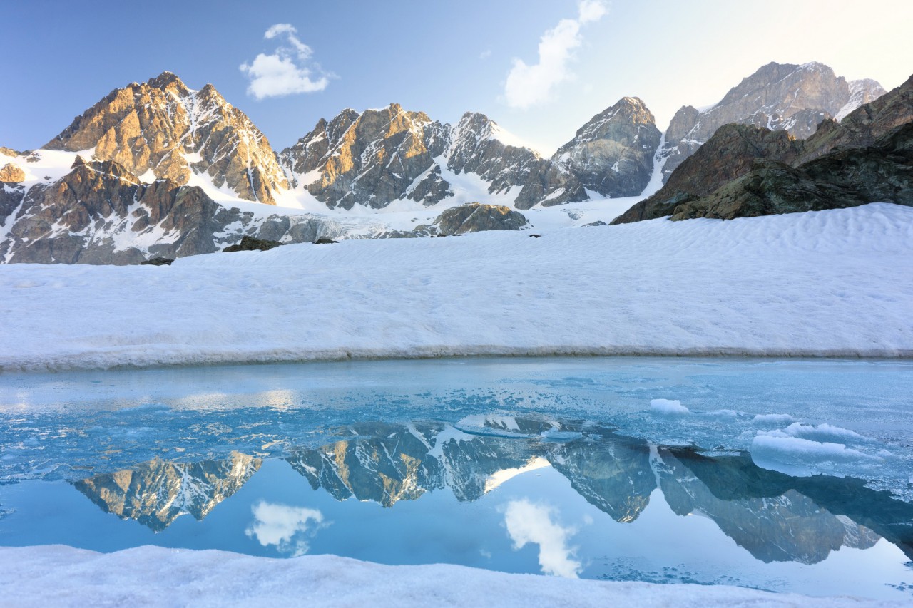 Ein halb zugefrorener, eisblauer Gebirgsee mit schneebedeckten Berggipfeln im Hintergrund