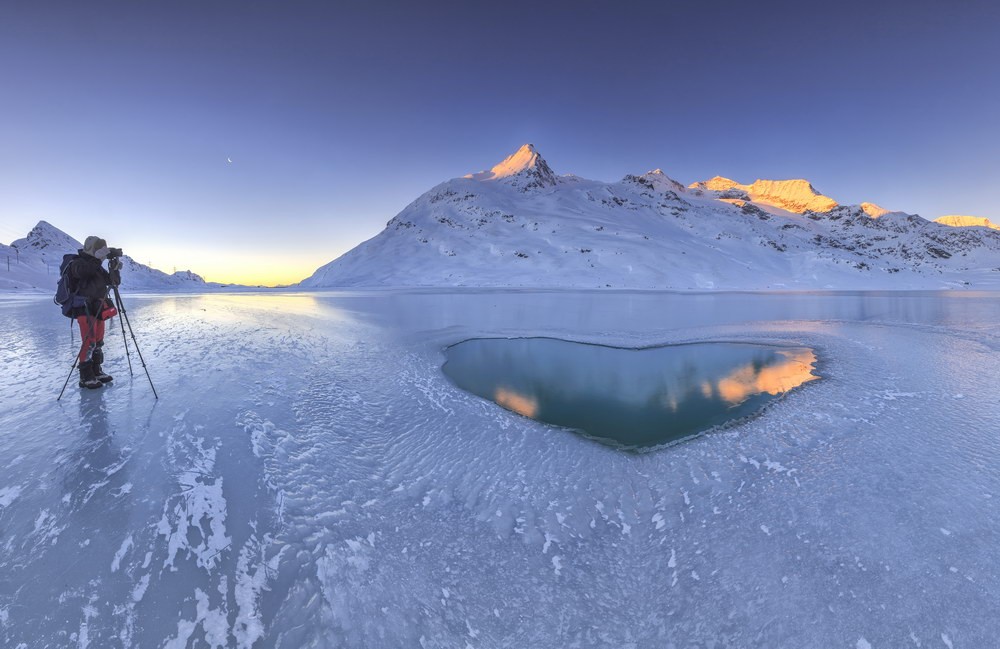 Fotograf fotografiert die ersten Sonnennstrahlen auf den beschneiten Gipfeln und einem Herz aus Wasser inmitten des zugeforenen Lago Bianco