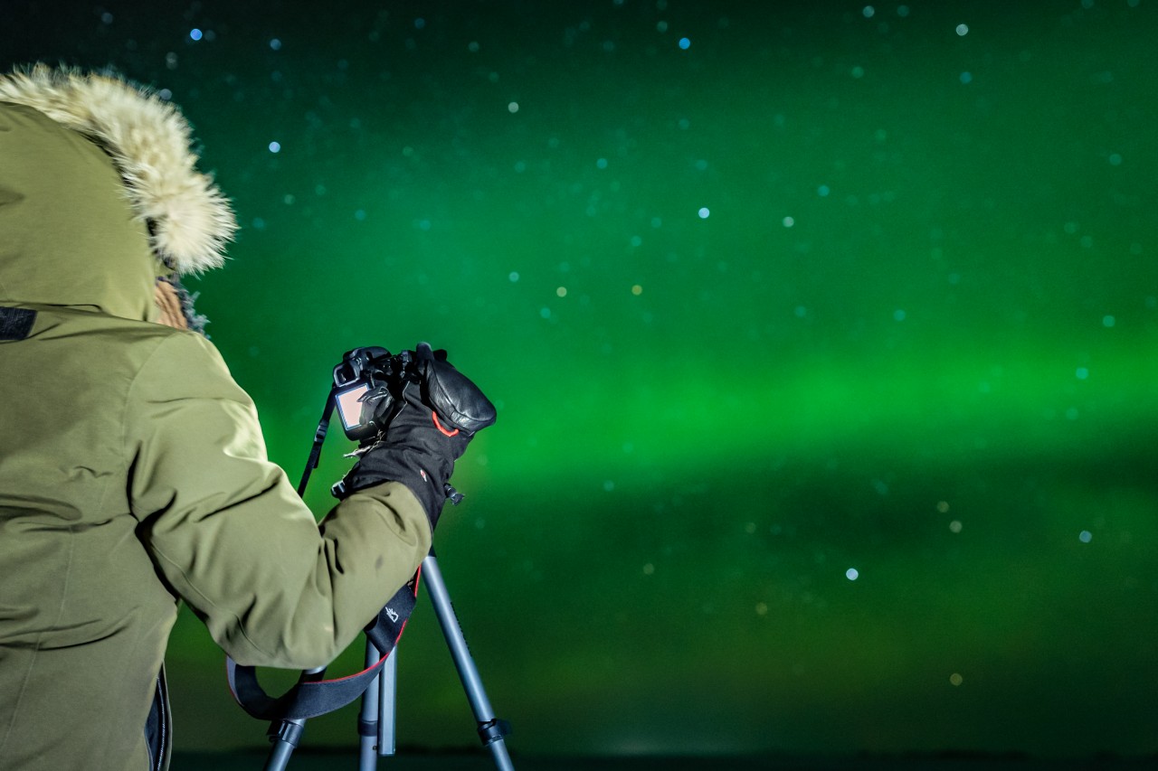 Une personne portant une épaisse veste d'hiver et des moufles, un appareil photo et un statif, photographie le ciel vert de la nuit