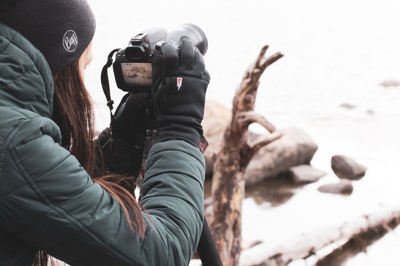 Photographer with gloves, jacket and hat takes a photo of a mountain backdrop
